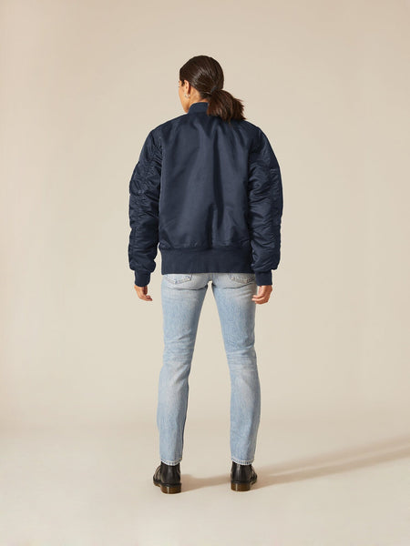 A person wearing an Alpha Industries MA-1 Battlewash Bomber Jacket in navy, accompanied by light blue jeans and black shoes, stands with their back to the camera.