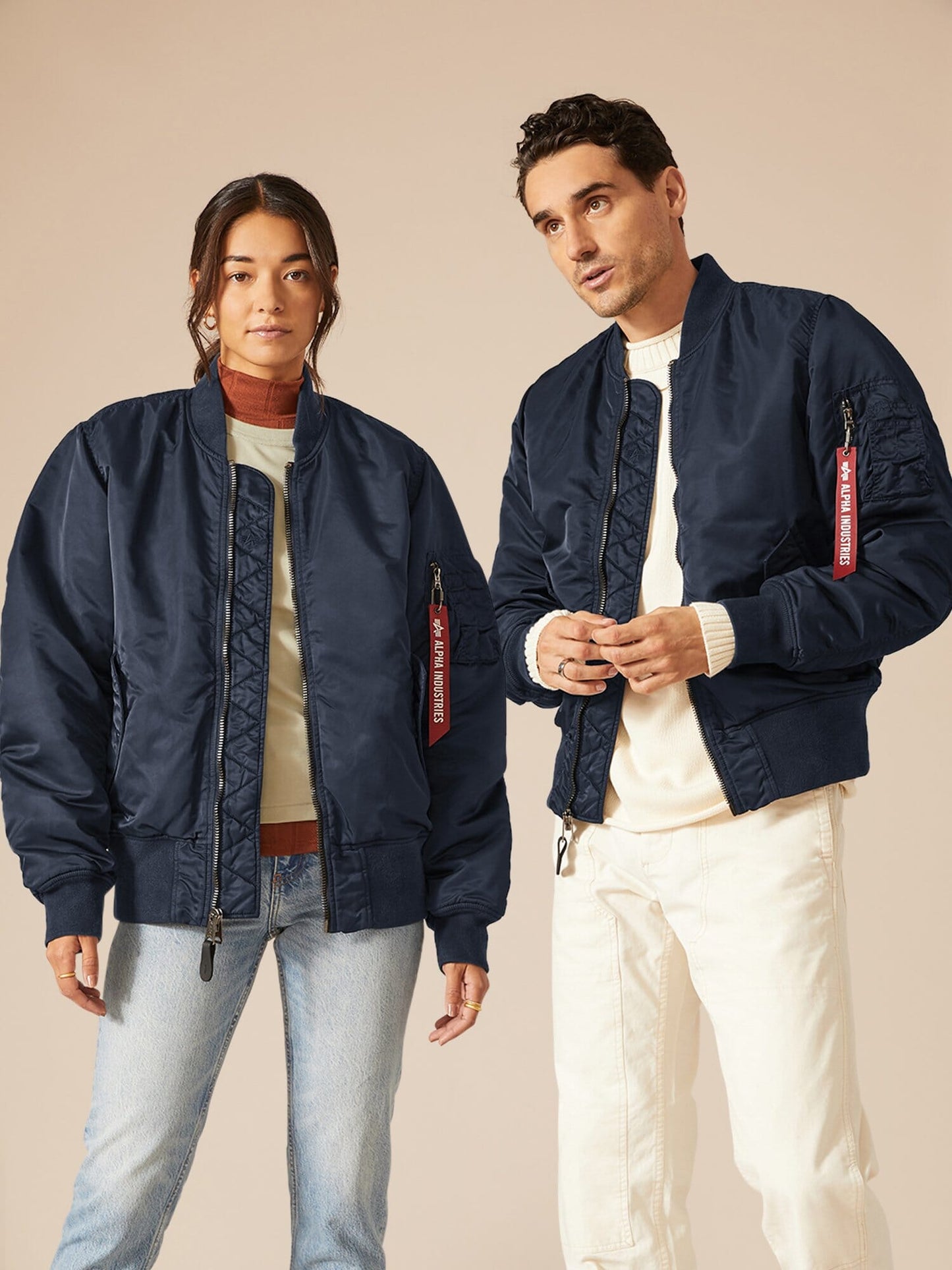 Two individuals don matching navy MA-1 Battlewash Bomber Jackets from Alpha Industries, one pairing it with light jeans and a beige sweater, the other with blue jeans and a t-shirt.