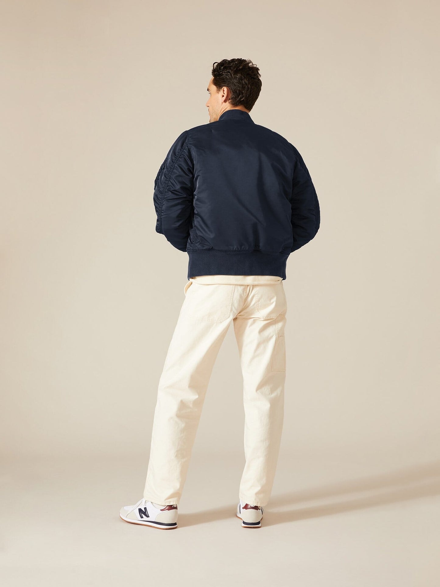 A person stands facing away, wearing an Alpha Industries, Inc. MA-1 Battlewash Bomber Jacket, beige pants, and white sneakers with red accents.