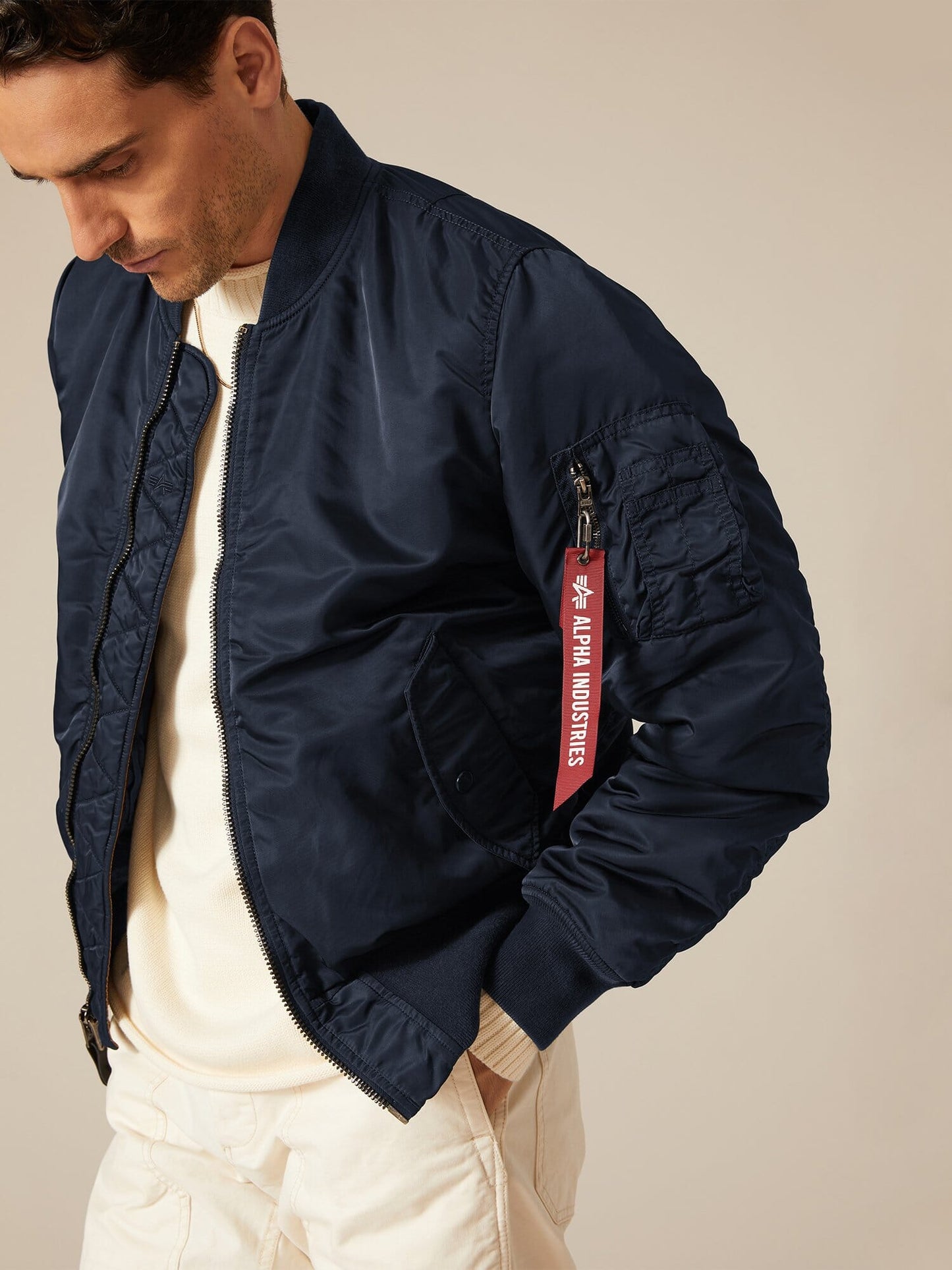 A person is wearing a navy MA-1 Battlewash Bomber Jacket by Alpha Industries, featuring a red Remove Before Flight tag on the sleeve, paired with a white shirt and pants.