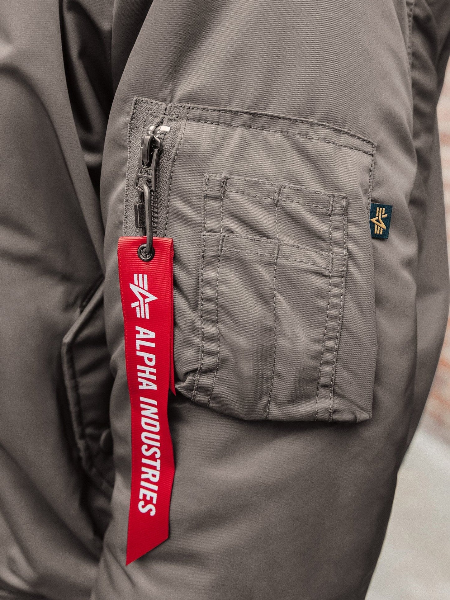 Side view of MA-1 Bomber Jacket (Heritage) in Gray. Red ribbon on utility pocket. By Alpha Industries.