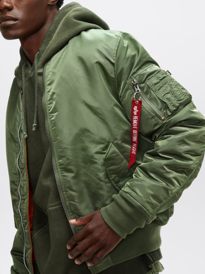 Sporting the Alpha Industries, Inc. MA-1 Bomber Jacket Slim Fit in green, featuring a red Remove Before Flight tag, and stylishly layered over a green hoodie.