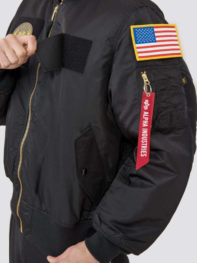Alpha Industries MA-1 Flex Bomber Jacket in black with U.S. flag patch, gold zipper, and red tag on sleeve.