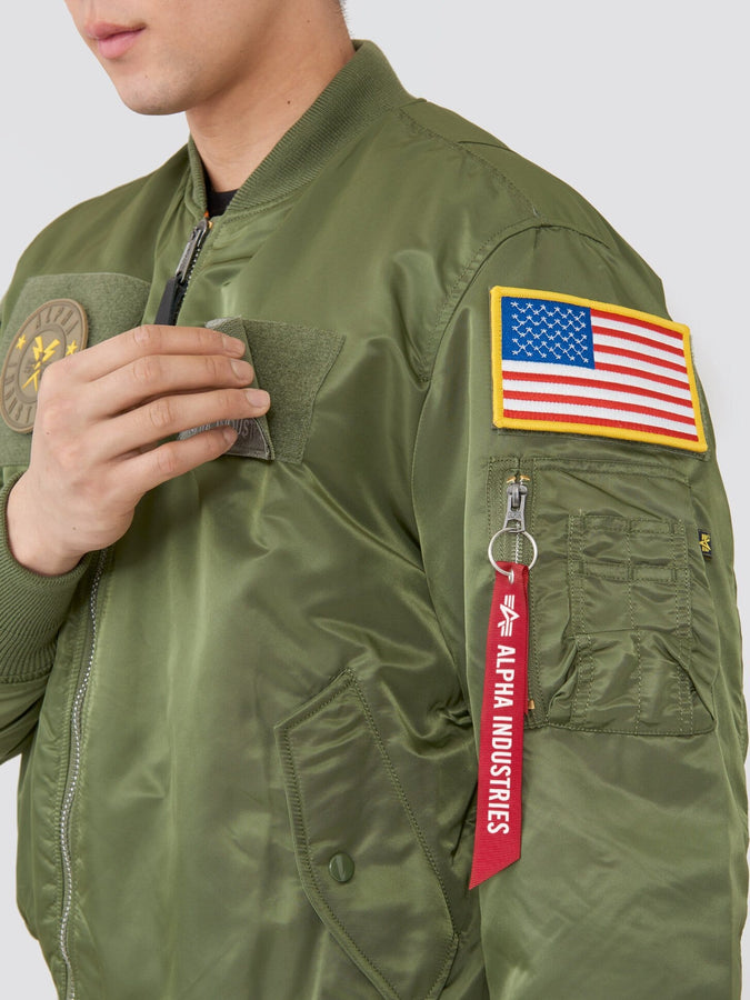 Alpha Industries MA-1 Flex Bomber Jacket in olive green with American flag patch, circular patch, and red Remove Before Flight tag on sleeve.