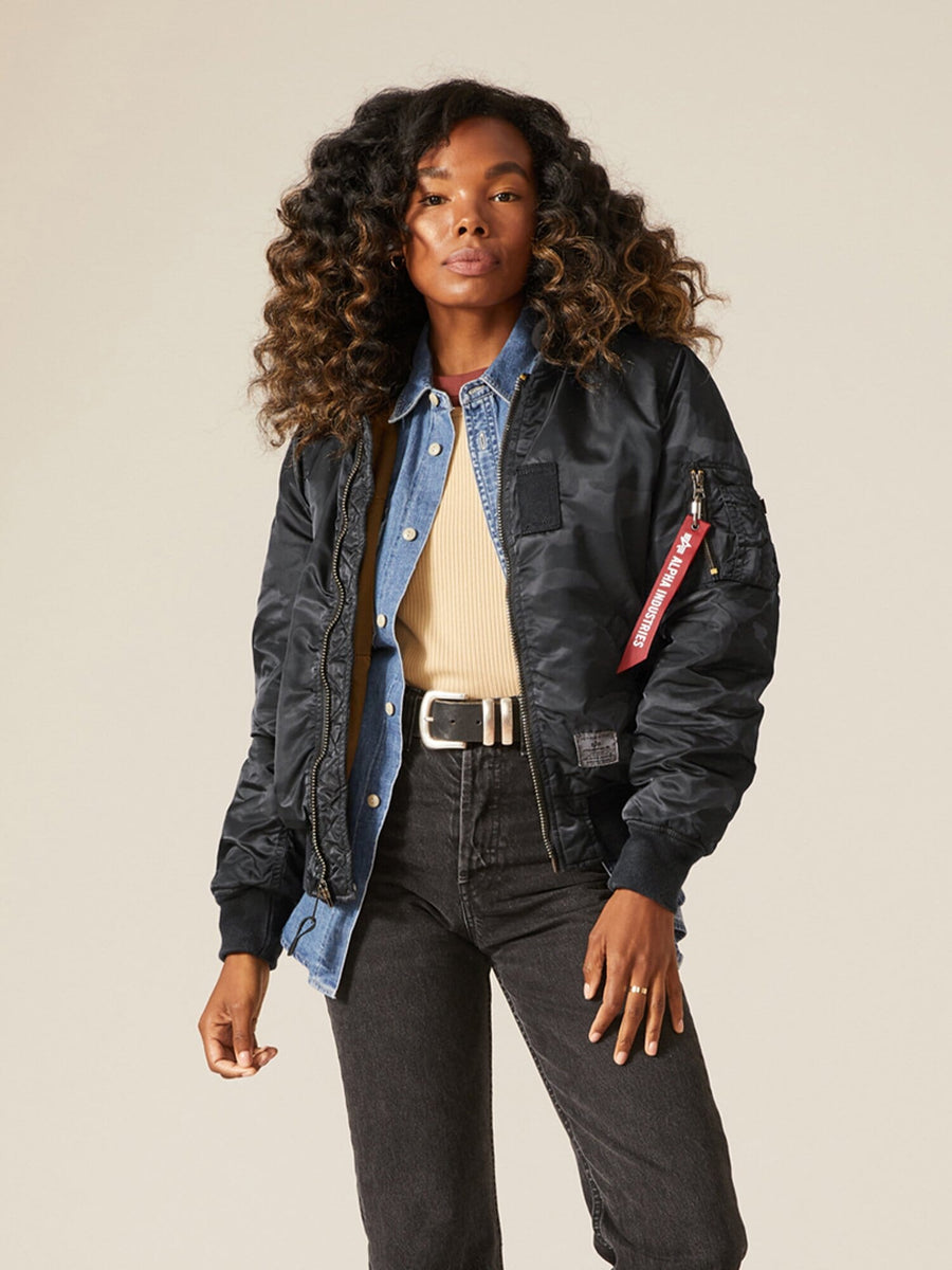 A person wearing an Alpha Industries MA-1 Hooded Battlewash Bomber Jacket in Black Woodland Camo over a denim jacket and beige shirt, paired with dark jeans.