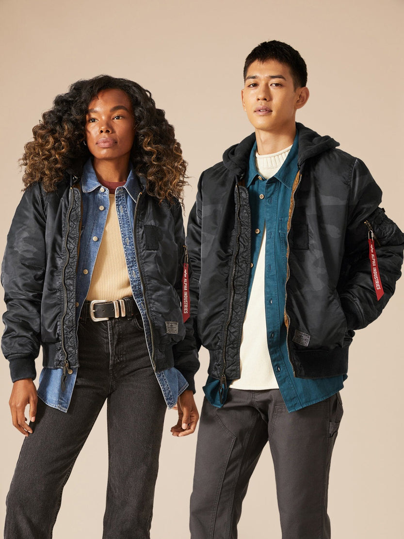 Two individuals wear denim shirts layered with Alpha Industries' MA-1 Hooded Battlewash Bomber Jackets. The person on the left flaunts long curly hair, while the one on the right has short hair. They seamlessly merge rugged style with timeless appeal.