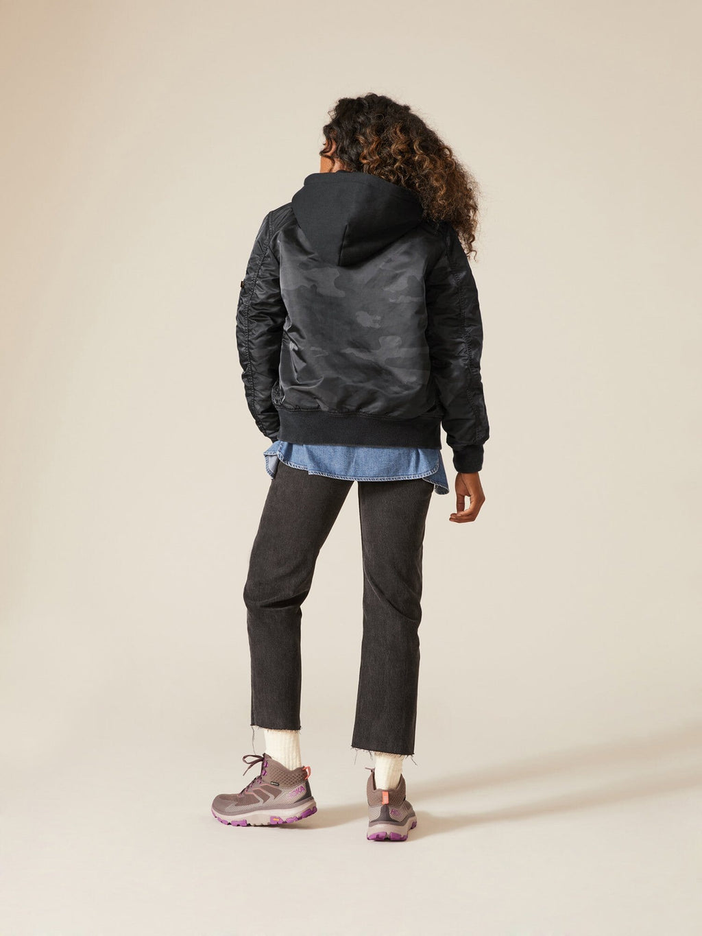 A person with curly hair faces away, wearing an Alpha Industries MA-1 Hooded Battlewash Bomber Jacket, a denim shirt, black pants, and pink sneakers.