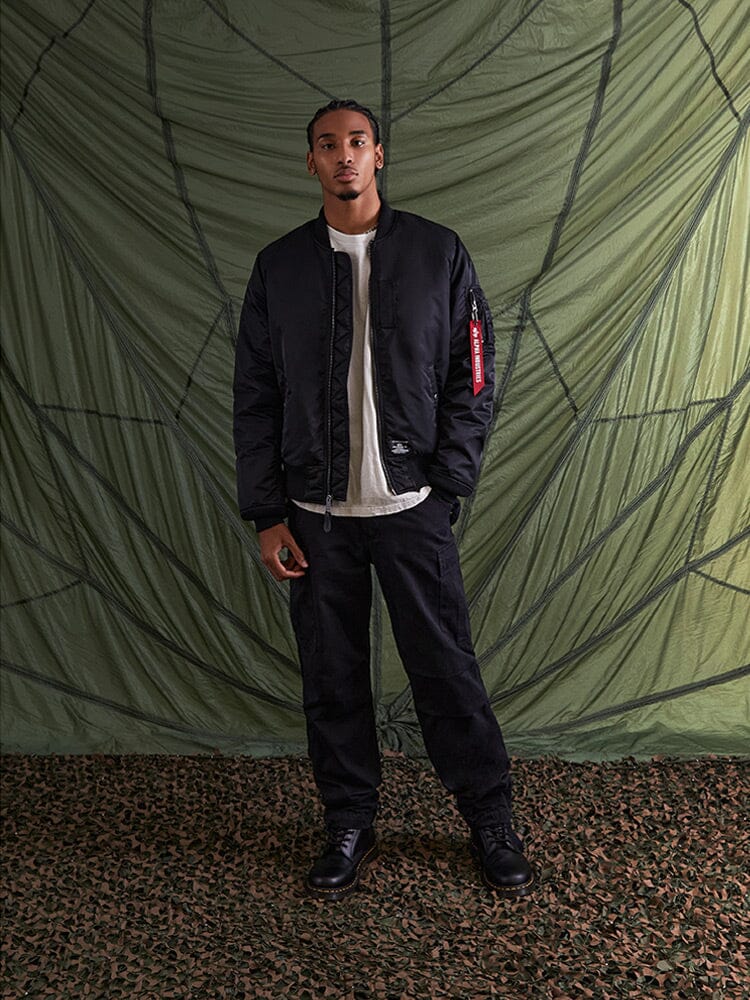 Alpha Industries MA-1 Mod Bomber Jacket in black with utility pocket, white t-shirt, black pants, and black boots.