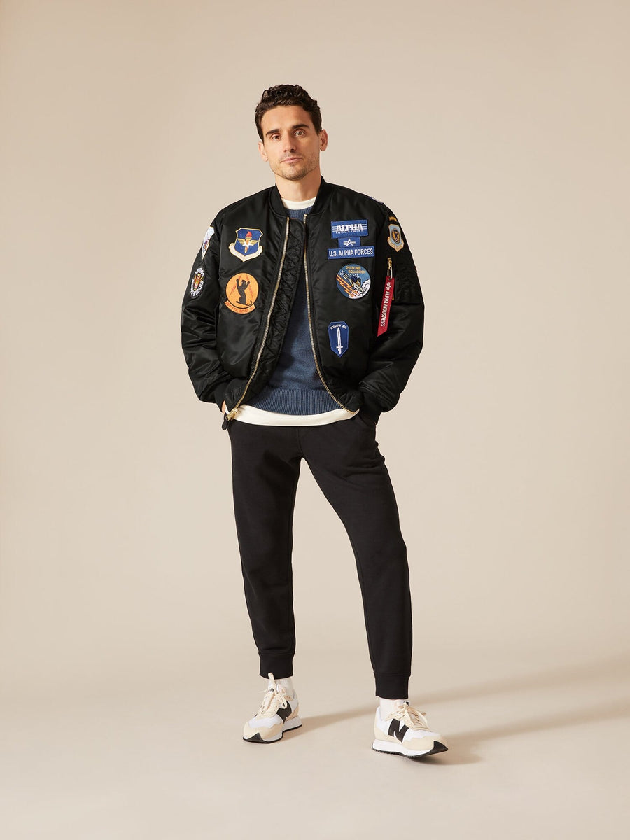 Alpha Industries MA-1 Squadron Bomber Jacket in black with colorful patches, blue shirt, black pants, and white sneakers.