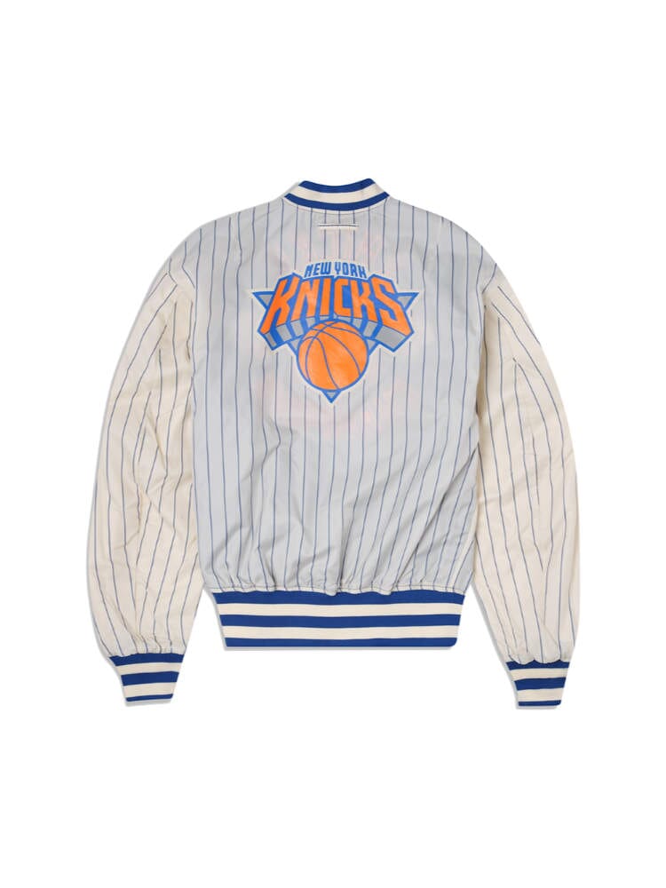 The New York Knicks x Alpha x New Era L-2B Bomber Jacket by Alpha Industries is a blue and white striped bomber jacket with the New York Knicks logo on the back, adorned with team patches.