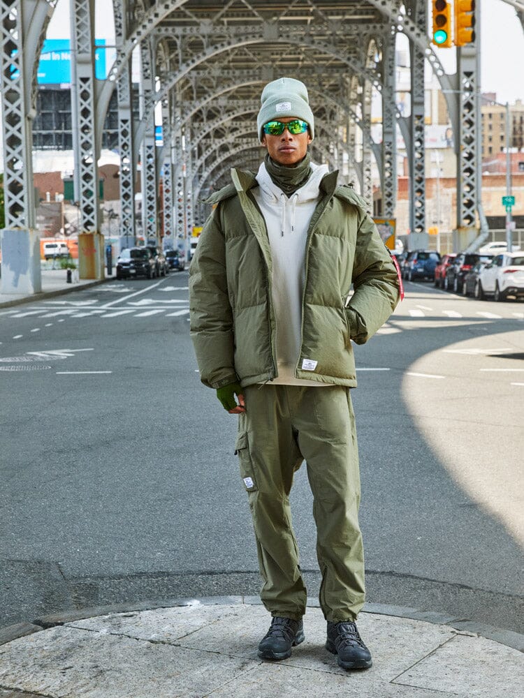 A person is standing under a steel bridge wearing the olive green Puffer Parka from Alpha Industries.