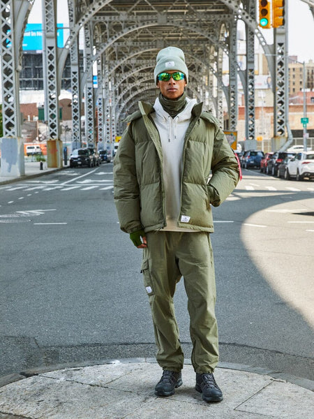 A person is standing under a steel bridge wearing the olive green Puffer Parka from Alpha Industries.
