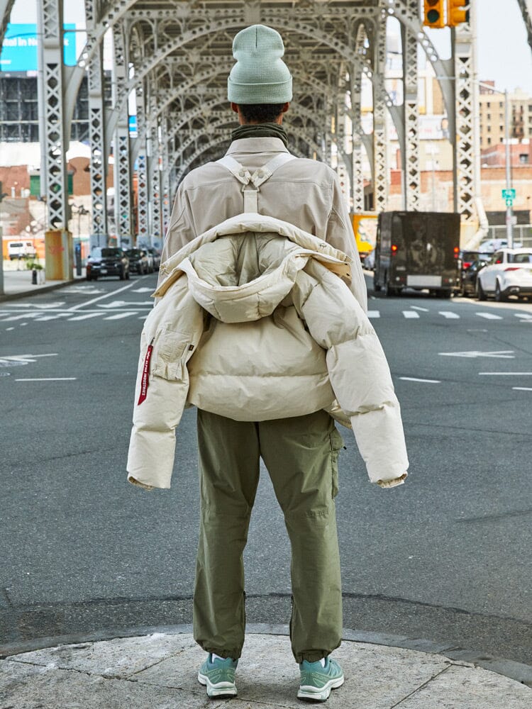 A person is standing under a steel bridge wearing the limestone Puffer Parka from Alpha Industries, showcasing the interior straps.