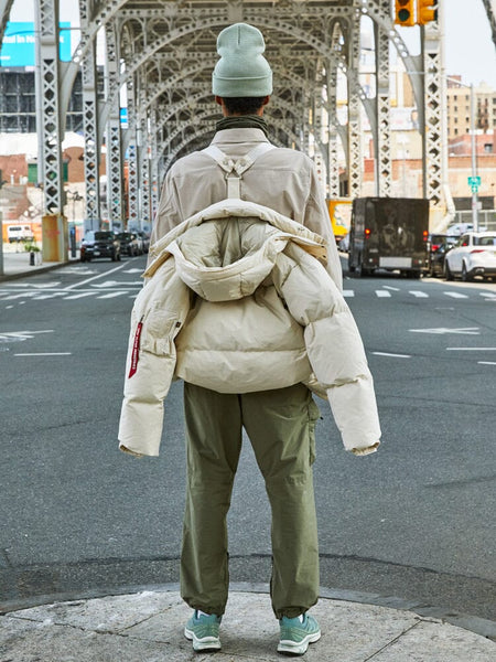 A person is standing under a steel bridge wearing the limestone Puffer Parka from Alpha Industries, showcasing the interior straps.