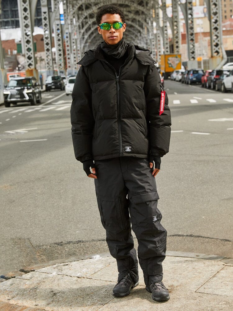 A person is standing under a steel bridge wearing the black Puffer Parka from Alpha Industries.