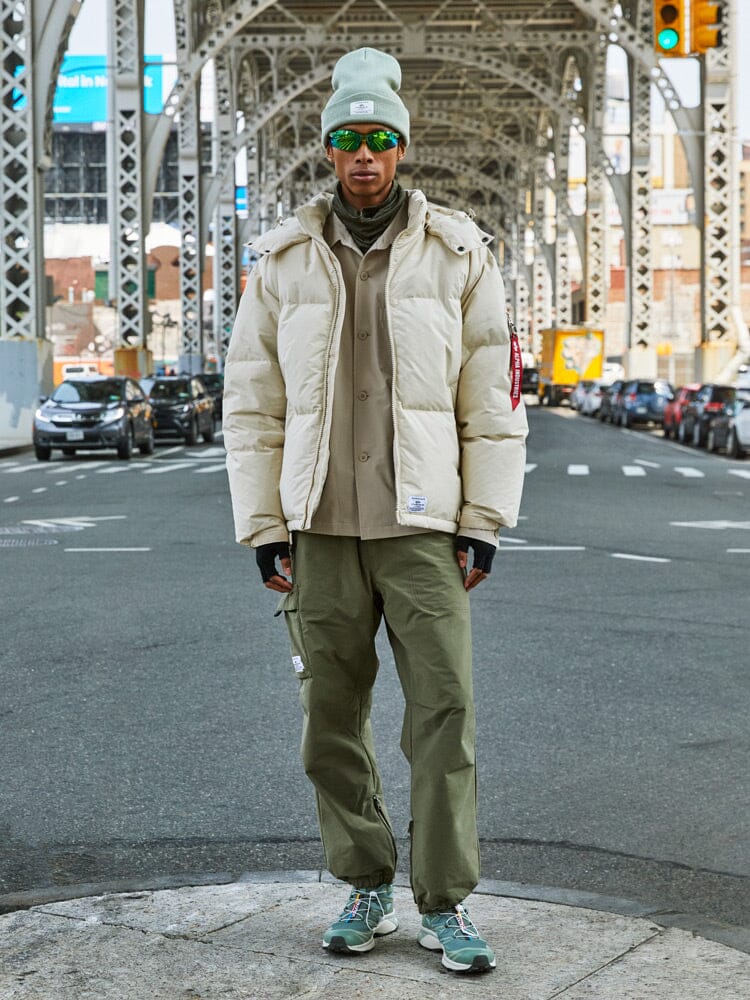A person dressed in a limestone Puffer Parka from Alpha Industries, as they stand on a city street under a bridge.