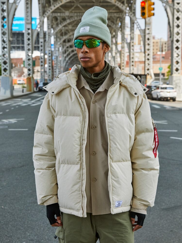 A person in an Alpha Industries Puffer Parka in limestone paired with reflective sunglasses stands under an urban bridge.
