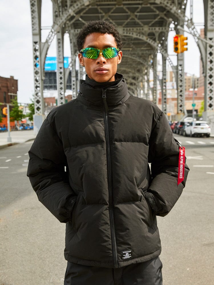 A person in an Alpha Industries Puffer Parka in black paired with reflective sunglasses stands under an urban bridge.