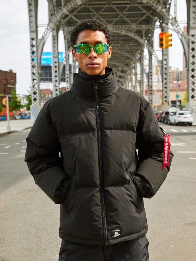 A person in an Alpha Industries Puffer Parka in black paired with reflective sunglasses stands under an urban bridge.