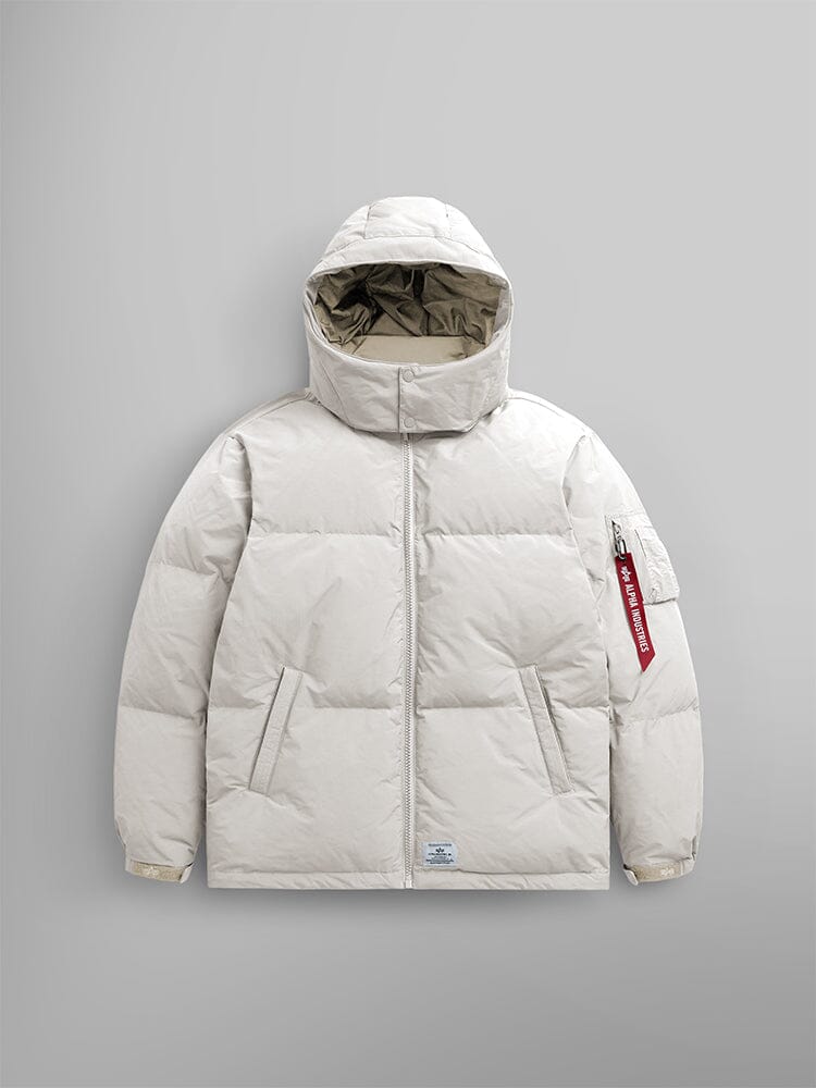 The limestone Puffer Parka by Alpha Industries is showcased with a removable hood and adjustable drawcords, along with a distinctive red tag on the sleeve.
