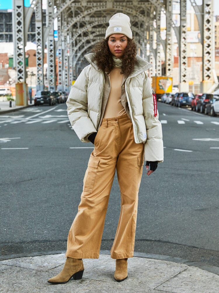 A person stands confidently on a city street under a bridge, wearing a beige ensemble featuring the water-resistant Sierra Short Puffer W by Alpha Industries, complemented with matching pants, beanie, and boots.