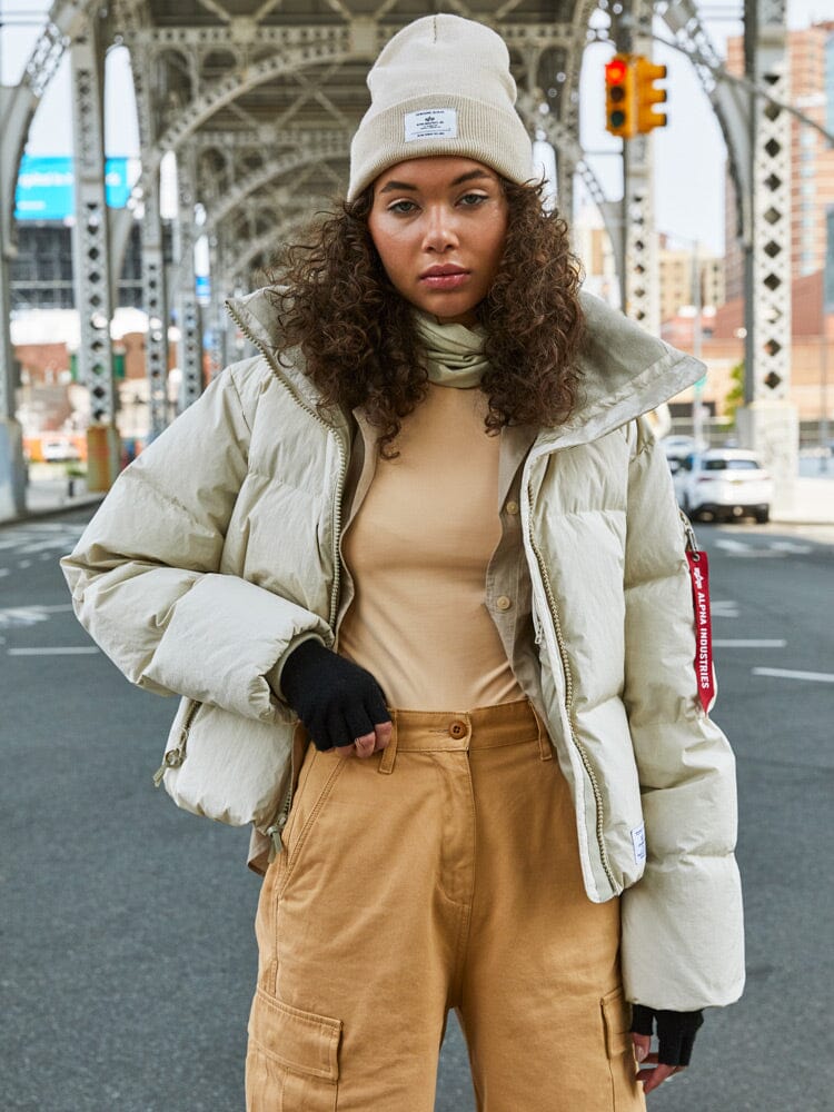 A person with curly hair is wearing the Sierra Short Puffer W, a water-resistant beige jacket by Alpha Industries, along with a beanie and tan pants while standing under an iron bridge in a city setting.