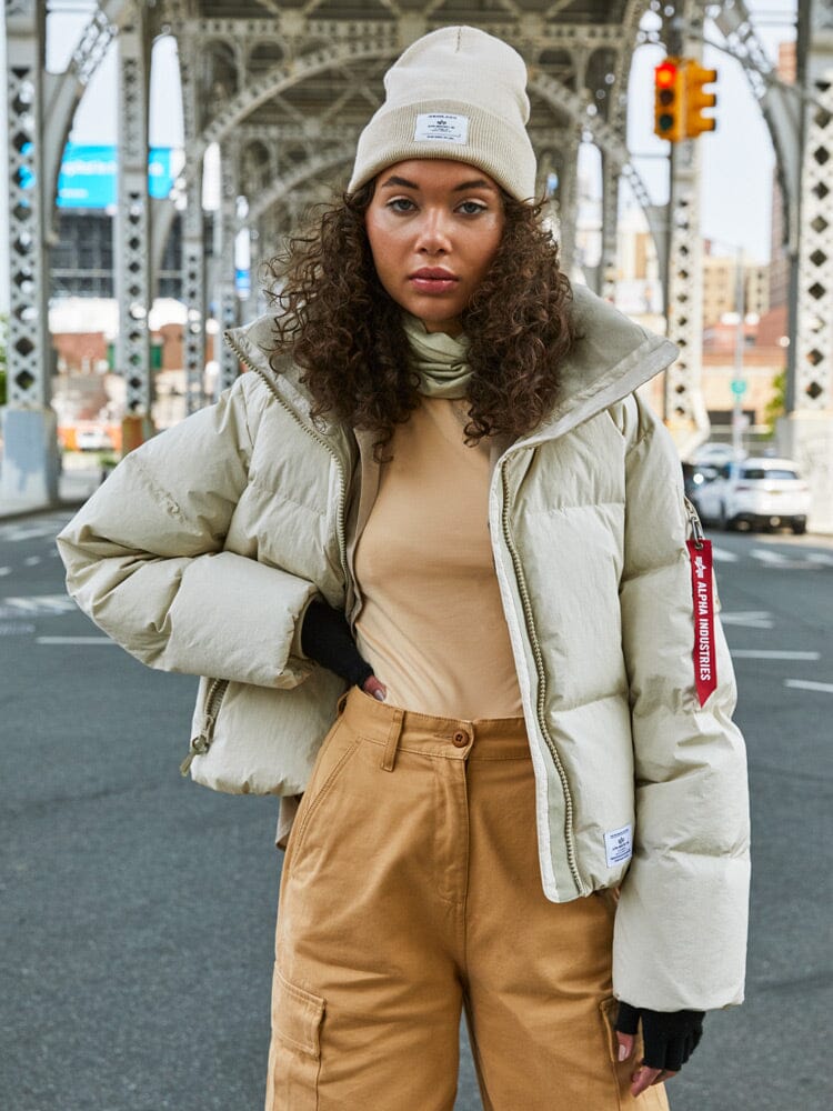 A person wearing a water-resistant, beige Sierra Short Puffer W parka by Alpha Industries, along with a beige beanie and tan pants, stands under a bridge in an urban setting.