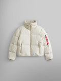 The Sierra Short Puffer W by Alpha Industries is a limestone-colored puffer parka with a high collar and water-resistant fabric. It includes two front zipper pockets and features a distinct red tag on the left sleeve.