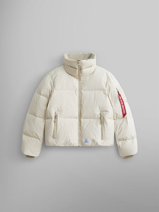 The Sierra Short Puffer W by Alpha Industries is a limestone-colored puffer parka with a high collar and water-resistant fabric. It includes two front zipper pockets and features a distinct red tag on the left sleeve.
